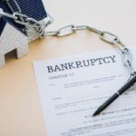 High angle view of house model with chain near pen and document with bankruptcy and chapter 13 lettering on beige High angle view of house model with chain near pen and document with bankruptcy and chapter 13 lettering on beige
